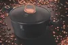 A black pot with rose gold accents sits on a dark surface scattered with rose gold-colored particles. Náhled obrázku produktu