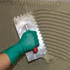 A gloved hand uses a notched trowel to spread adhesive on a wall. Anteprima dell’immagine prodotto