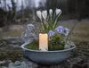 A decorative outdoor arrangement with a candle, white crocuses, and purple pansies in a grey bowl, placed on a mossy stone surface. Anteprima dell’immagine prodotto