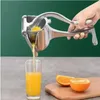 A hand is using a manual aluminum juicer to squeeze orange juice into a glass. Oranges are visible on a cutting board. Produktbild-Vorschau