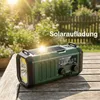 A green emergency radio with a solar panel on top sits on a wooden table outdoors, with a garden and fence in the background. Produktbild-Vorschau