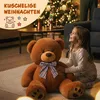 A young girl sits on the floor next to a large teddy bear in a living room decorated for Christmas. Anteprima dell’immagine prodotto