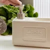 A hand is pressing down on a soap dispenser next to a scrub brush, with green leafy vegetables in the background. Náhľad obrázku produktu