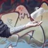 A person's tattooed arm and hand are shown with a white Bluetooth speaker levitating above their palm, against a graffiti-covered wall. Anteprima dell’immagine prodotto