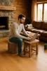 A man is working on a laptop while sitting on a log-shaped pouf in a cozy living room with a fireplace and leather sofa. Podgląd zdjęcia produktu