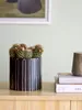 A black ceramic pot with white vertical stripes containing a cluster of small cacti with pink flowers sits on a wooden surface next to a stack of books. Produktbild-Vorschau