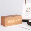 A wooden digital alarm clock with a wireless charging pad on top is displayed on a white surface next to a calendar. Produktbild-Vorschau