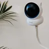 A white baby monitor camera is mounted on a flexible arm against a light-colored wall, with a plant leaf in the foreground. Anteprima dell’immagine prodotto