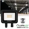 A black LED floodlight is shown in the foreground, with a blurred background of a warehouse interior illuminated by multiple ceiling lights. Voir les images du produit
