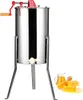 A stainless steel honey extractor with a red crank and wooden handle, standing on metal legs. Next to it are a pitcher of honey, a honey dipper, and honeycomb. Podgląd zdjęcia produktu