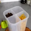 A clear plastic food storage container with multiple compartments, filled with coffee beans and pasta, sits on a grey surface next to a green pear. Produktbild-Vorschau