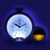 A close-up, illuminated view of a children's alarm clock and sleep trainer, set against a dark blue background. Produktbild-Vorschau