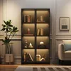 A black glass display cabinet with LED lighting, filled with various decorative items and books, placed in a room next to a potted plant and a sofa. Produktbild-Vorschau