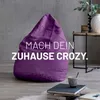 A purple beanbag chair is placed in a modern living room setting, with a side table and books nearby. Produktbild-Vorschau