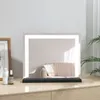 A lighted makeup mirror sits on a wooden dresser, reflecting a couch with pillows.