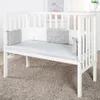 A white crib with a light gray mattress and a patterned bumper pad. Produktbild-Vorschau
