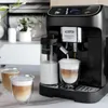 A black DeLonghi Magnifica Plus coffee machine is shown making a latte, with two finished lattes in the foreground. Produktbild-Vorschau