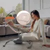 A woman is sitting on the floor next to a baby in a bouncer, looking at the baby and smiling. The scene is set in a living room with a sofa and large windows in the background. Produktbild-Vorschau