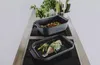 Two black roasting pans are placed on a modern stovetop, one containing a prepared fish and vegetables, the other containing roasted potatoes. Náhľad obrázku produktu
