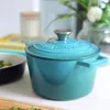 A teal Dutch oven sits on a white surface, with a blurred background of books, a candle, and greenery. Produktbild-Vorschau