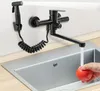 A black kitchen faucet with a side sprayer is mounted on a wall above a silver sink. A hand is holding a red tomato under the running water from the faucet. Náhled obrázku produktu
