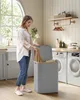 A woman is putting laundry into a grey laundry basket with a bamboo lid in a laundry room. Náhled obrázku produktu