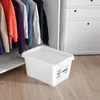 A white storage box with a lid sits on a wooden floor in front of a closet with clothes hanging inside. Produktbild-Vorschau