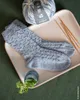 A pair of knitted socks, a ball of yarn, and knitting needles are arranged on a pink tray, with a small plant in a blue vase in the foreground. Produktbild-Vorschau