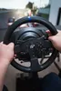 A person's hands are holding a racing steering wheel, playing a racing game on a screen in the background. Produktbild-Vorschau