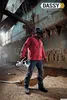 A worker in a red jacket and blue jeans stands in an industrial setting, holding a piece of equipment. Produktbild-Vorschau