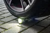A flashlight is placed on a paved surface under a car tire, with its light illuminating the ground. Produktbild-Vorschau