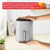 A hand is shown placing a compact air fryer on a wooden countertop, with books and a plant in the background. Produktbild-Vorschau