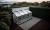 A greenhouse attached to a dark-colored building, set in a garden with trees and a hedge. Náhled obrázku produktu