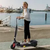 A young girl is riding an electric scooter by a harbor with boats in the background. Produktbild-Vorschau