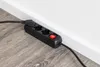 A black power strip with three outlets and a red on/off switch is placed on a wooden floor next to a white baseboard and wall. Produktbild-Vorschau