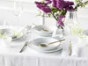 A table setting with white plates, gold cutlery, and a vase of purple lilacs. Produktbild-Vorschau