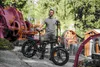 A man stands with his electric bicycle in an outdoor setting with industrial machinery and trees in the background. Produktbild-Vorschau