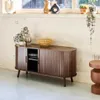 A wooden media console with ribbed doors is placed in a room with a window and several decorative items on top and next to it. Anteprima dell’immagine prodotto