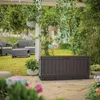 A dark brown storage box is placed on a stone patio in a garden setting, with outdoor furniture and plants in the background. Podgląd zdjęcia produktu