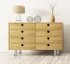 A wooden dresser with eight drawers, supported by four silver cylindrical legs, is shown against a white wall and on a wooden floor. Decorative items, including a lamp, two wire spheres, and a vase with branches, are placed on top of the dresser. Produktbild-Vorschau