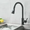 A black kitchen faucet with a pull-down sprayer is shown in use, with water flowing into a stainless steel sink. A bowl of produce is visible to the left. Produktbild-Vorschau