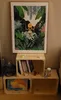 A framed artwork of a jungle scene with a person on a swing is displayed above a set of wooden crates. The crates are stacked and contain books, a small drawing, and a bottle. Produktbild-Vorschau