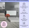 A white electric heater is mounted on a gray wall in a living room setting, with a sofa and a floor lamp. Produktbild-Vorschau