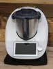 A white kitchen appliance with a black lid and handle, placed on a black base, with a wooden background. Produktbild-Vorschau