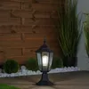 A black vintage-style outdoor lantern is placed on a paved patio in front of a wooden fence and some greenery. Produktbild-Vorschau