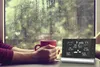 A person holding a mug of coffee in front of a window on a rainy day, with a weather station on the table. Podgląd zdjęcia produktu