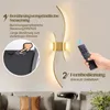 A modern gold wall lamp with two curved light elements is shown mounted on a wall above a sofa. The image highlights two control methods: touch control on the lamp itself and a remote control. Text overlays describe these features. Voir les images du produit