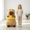 A young girl stands next to a large capybara plush toy in a brightly lit room. Anteprima dell’immagine prodotto