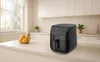 A black air fryer sits on a kitchen counter next to a bowl of fruit, with kitchen cabinets and a window in the background. Produktbild-Vorschau