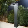 A modern, dark gray solar bollard light stands on a paved walkway next to lush green bushes and bamboo plants at dusk. Produktbild-Vorschau